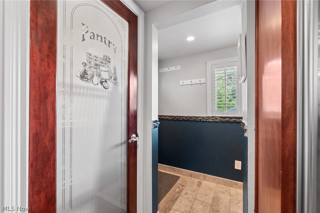 Pantry doorway with a decorative glass door, wall hooks, shuttered window, and tiled floor.