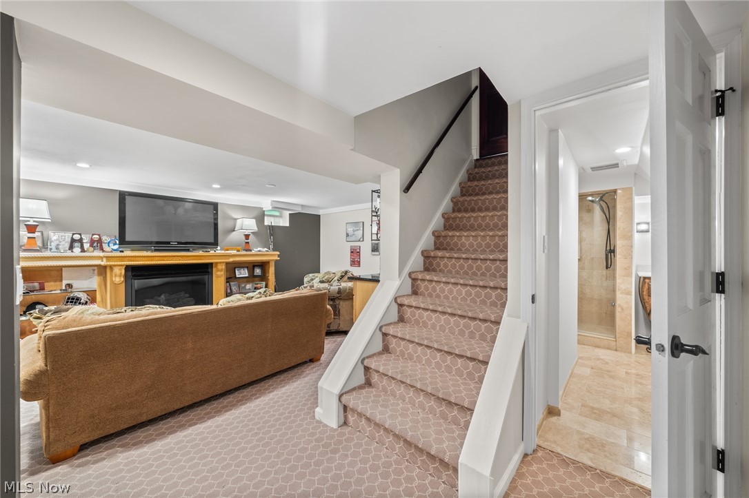 Furnished basement family room with wood fireplace mantel, carpeted stairs, and adjoining tiled bathroom.