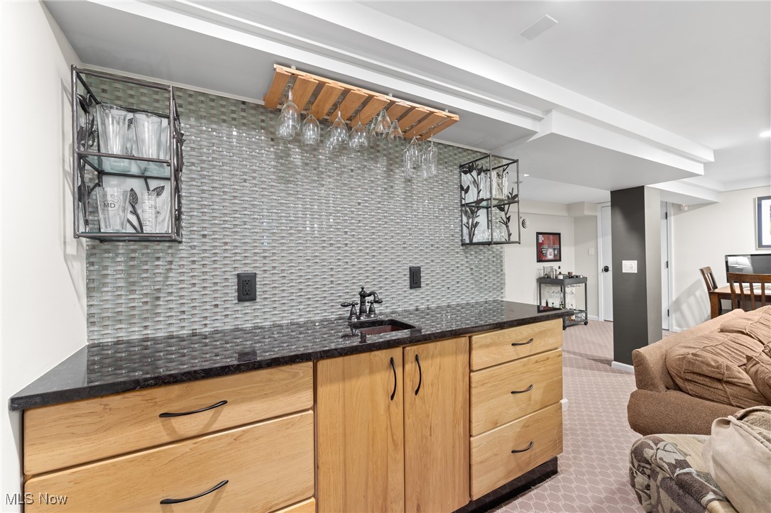Wet bar with light wood cabinets, textured gray backsplash, and hanging glass rack beside a seating area.