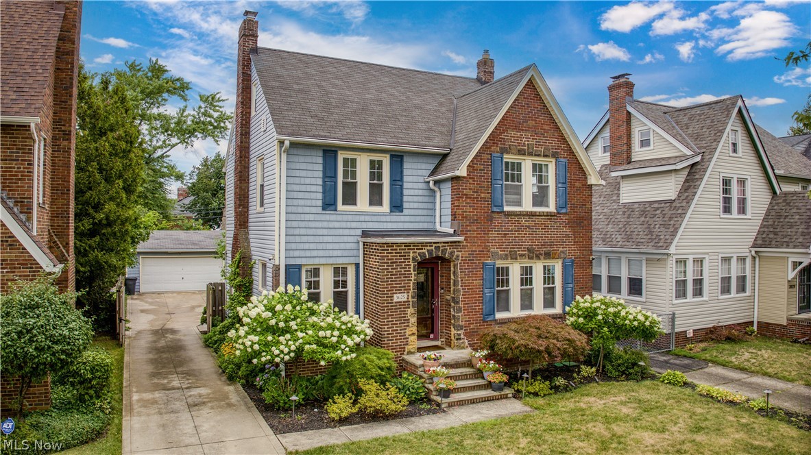 Two-story blue-gray and brick home with blue shutters, flowering shrubs, and a driveway leading to a garage.