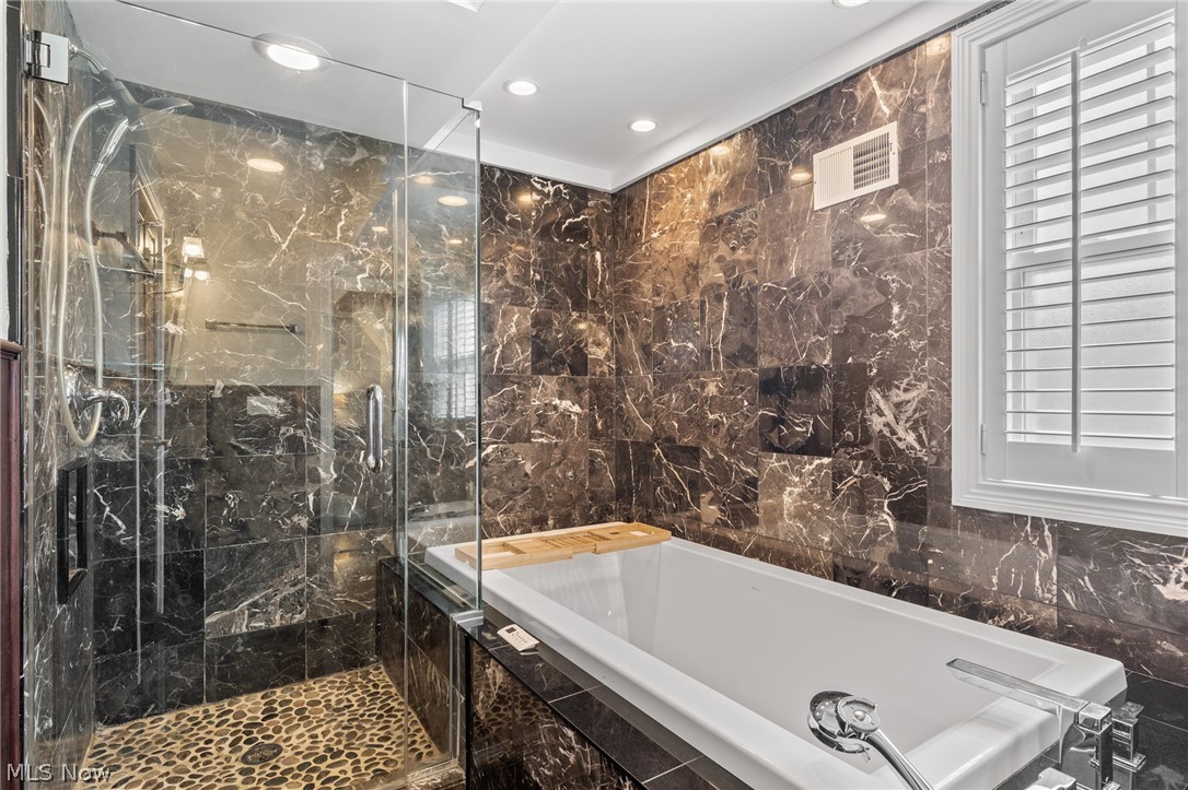 Bathroom with dark marble walls, glass shower with pebble floor, white soaking tub, and shuttered window.