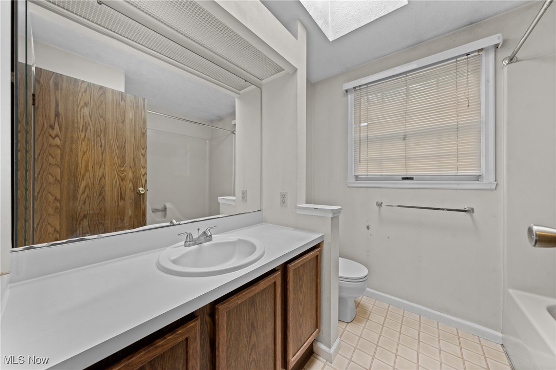 Bathroom with wood-tone vanity, large mirror, toilet, window with blinds, and a partially visible bathtub.