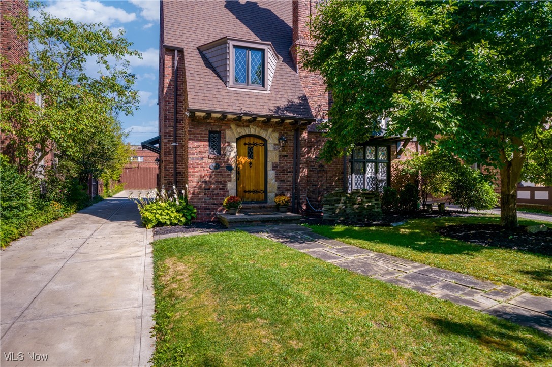 Brick home with a yellow front door, steep shingled roof, paved driveway, and mature trees.