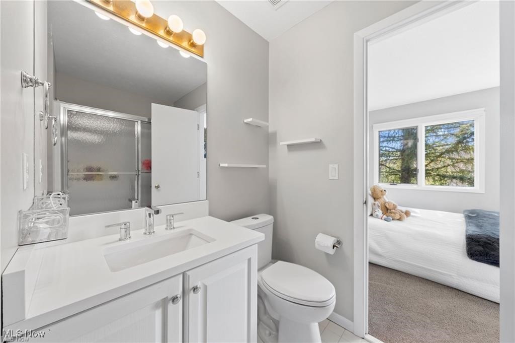 Bathroom with a white vanity and glass shower, opening onto a bedroom with a window-side bed.