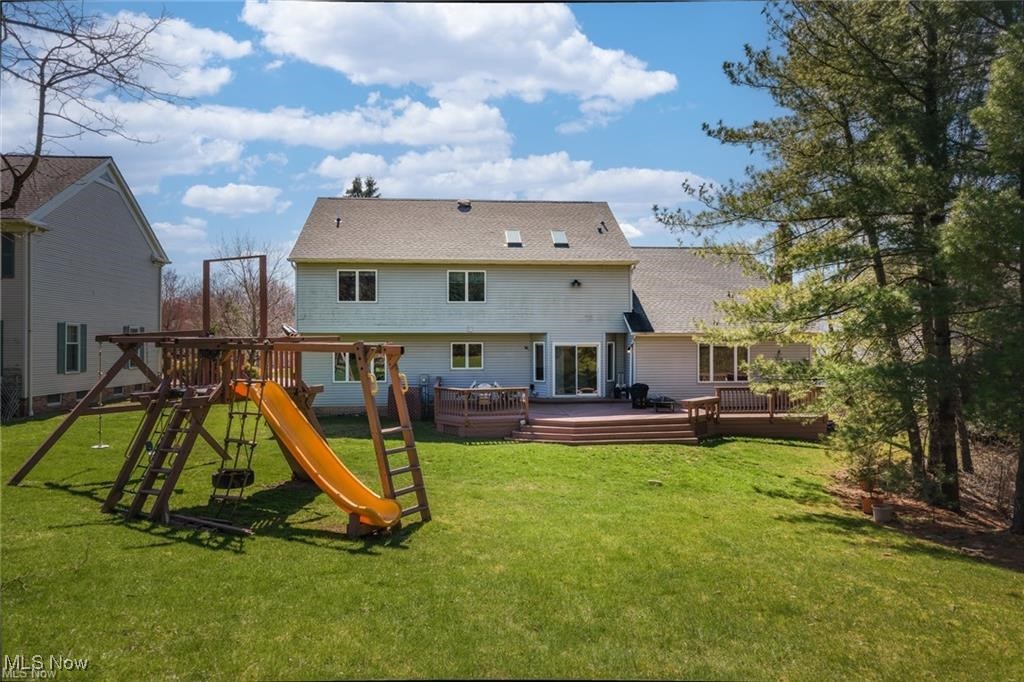 Rear of a gray two-story home with a wooden deck, broad lawn, and playset with an orange slide.
