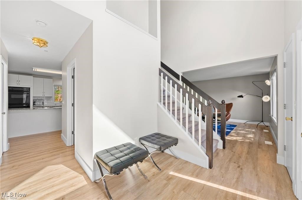 Bright foyer with a dark-railed staircase, two gray ottomans, and an adjoining kitchen.