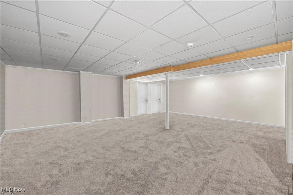 Empty carpeted basement recreation room with a drop ceiling, wood-trimmed beam, and support column.