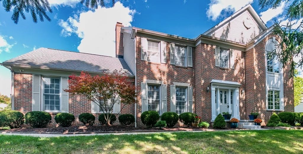 Two-story brick house with white trim, a gabled entrance, landscaped shrubs, and a green lawn.