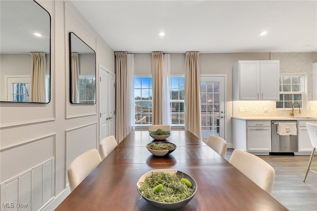Dining area with a long wooden table, cream chairs, tall curtained windows, and an adjoining white kitchen.