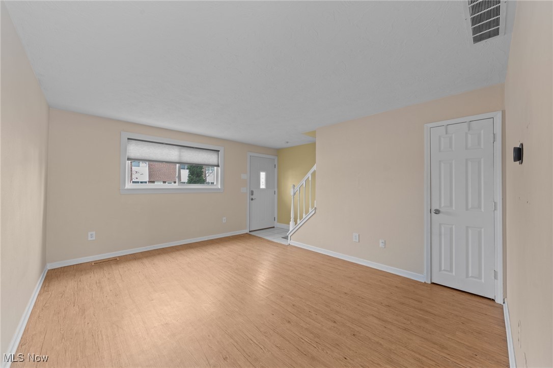 Empty room with light wood flooring, a wide window, front door, and staircase.