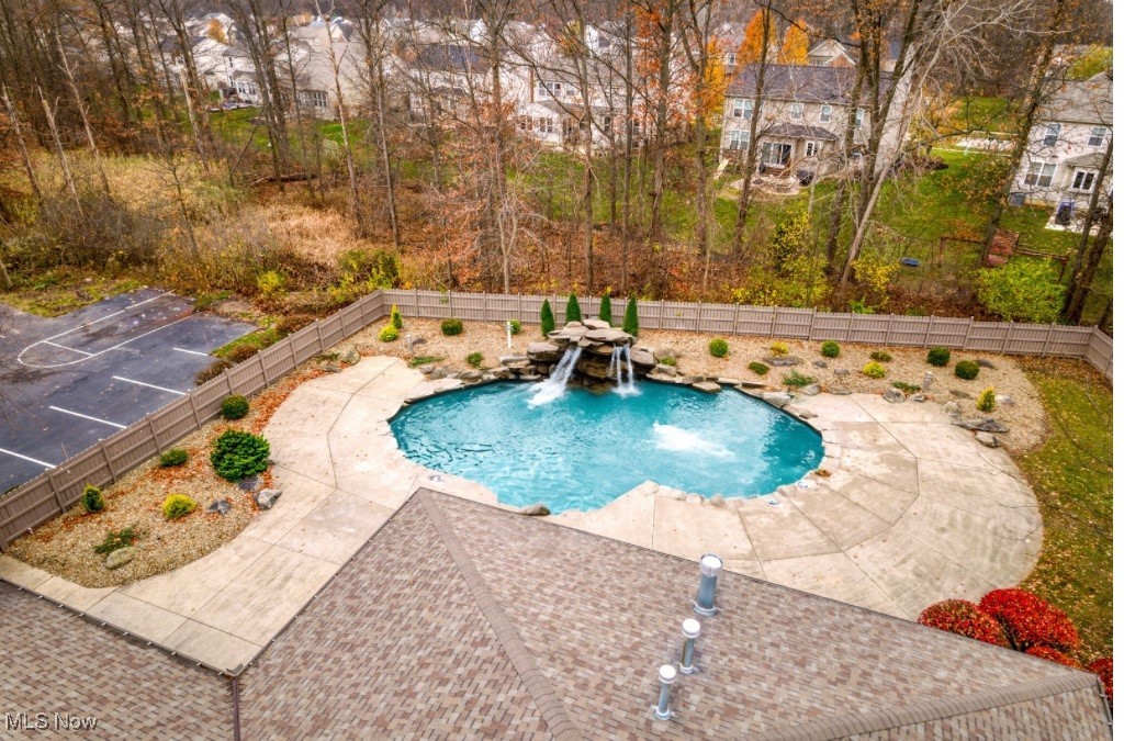 Backyard swimming pool with a rock waterfall, broad patio, and autumn trees beyond the fence.