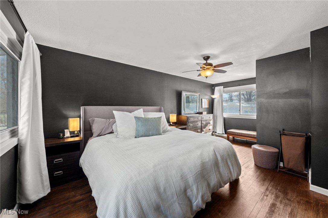 Furnished bedroom with a white bed, charcoal walls, hardwood floors, and large curtained windows.