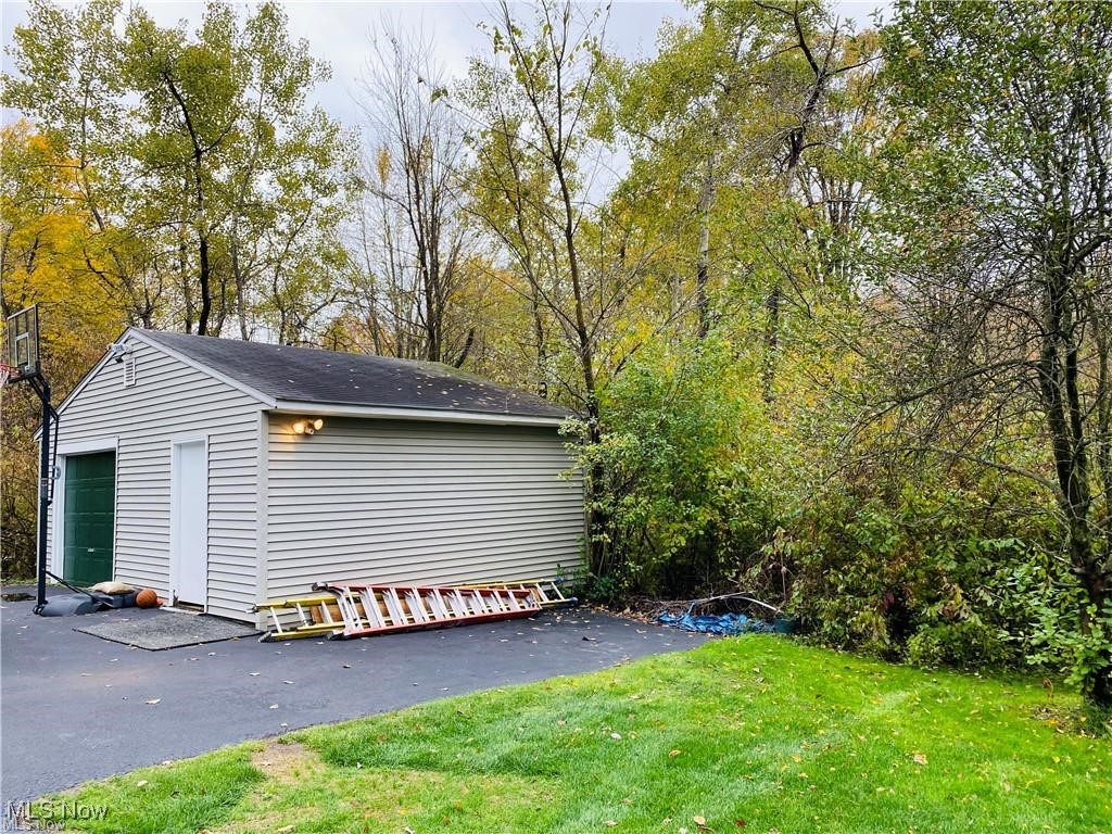 Detached garage beside a paved driveway, with a basketball hoop and wooded autumn backdrop.