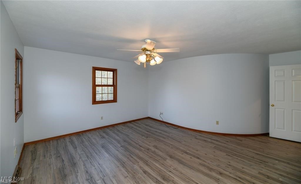 Empty room with gray wood-look flooring, two wood-trimmed windows, and a ceiling fan with lights.