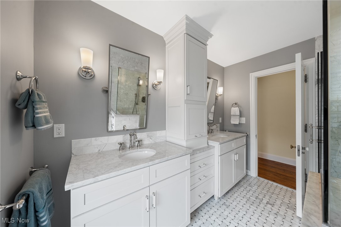 Bathroom with a marble double vanity, white cabinetry, patterned tile floor, and glass-enclosed shower.
