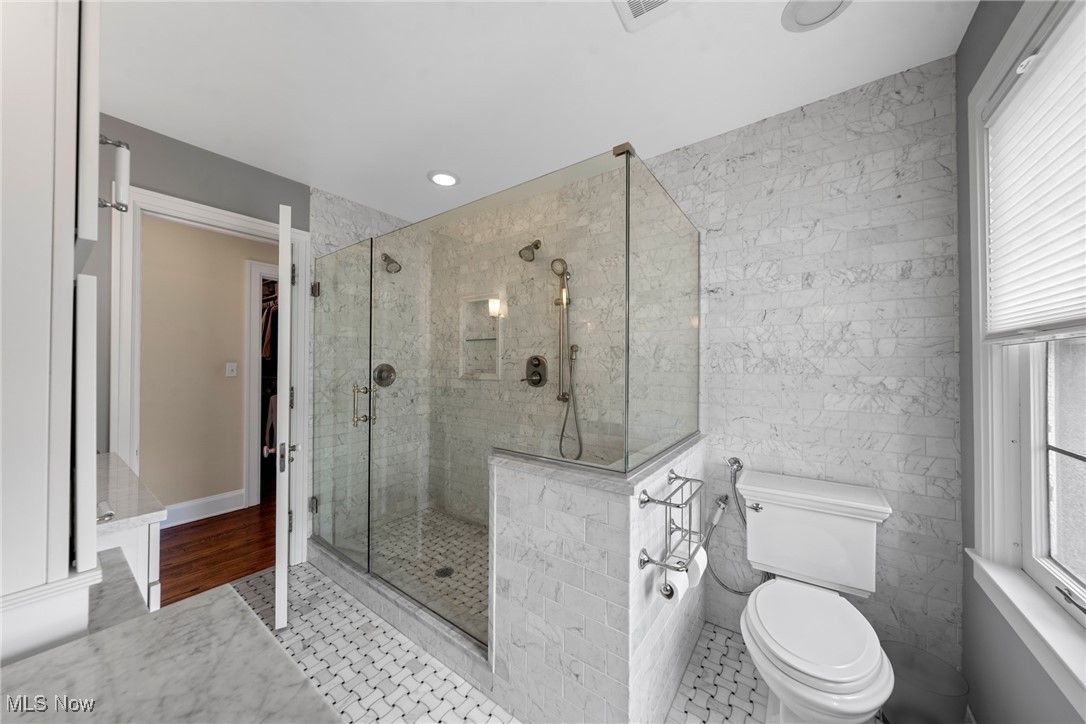 Marble-tiled bathroom with a glass walk-in shower, white toilet, and window with blinds.