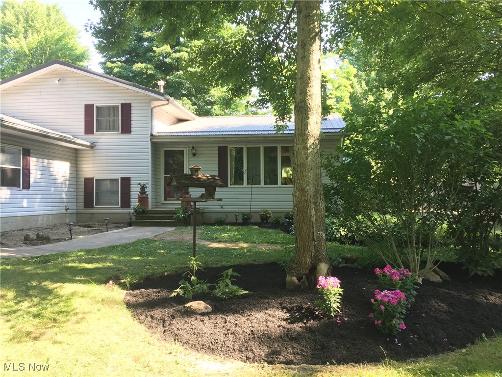 Light-sided house with burgundy shutters, a small patio, and a flower bed beneath mature trees.