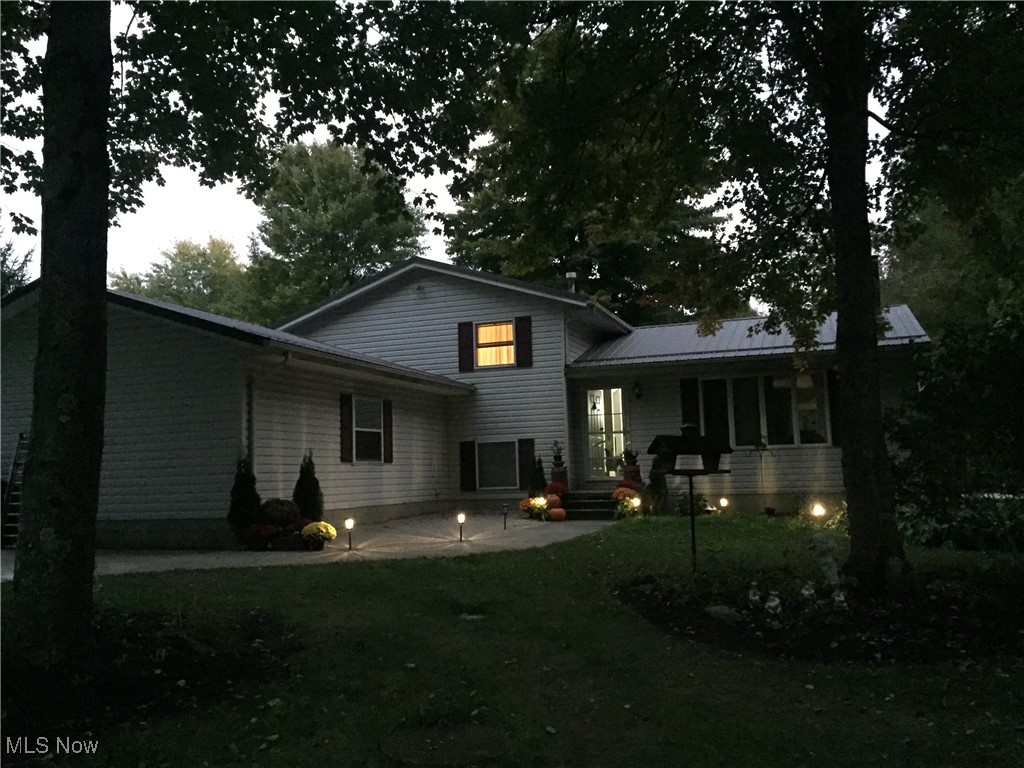 Gray-sided house at dusk, with a covered porch and illuminated windows beneath mature trees.