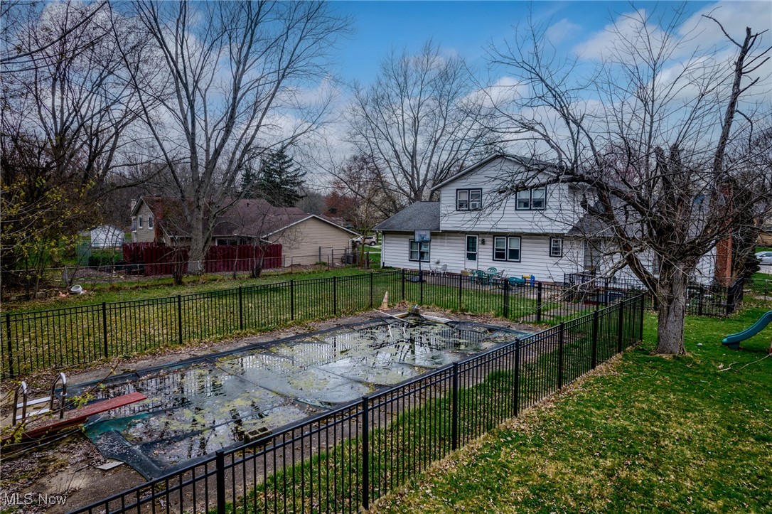 Backyard pool with standing water and leaves, enclosed by a black fence behind a two-story house.