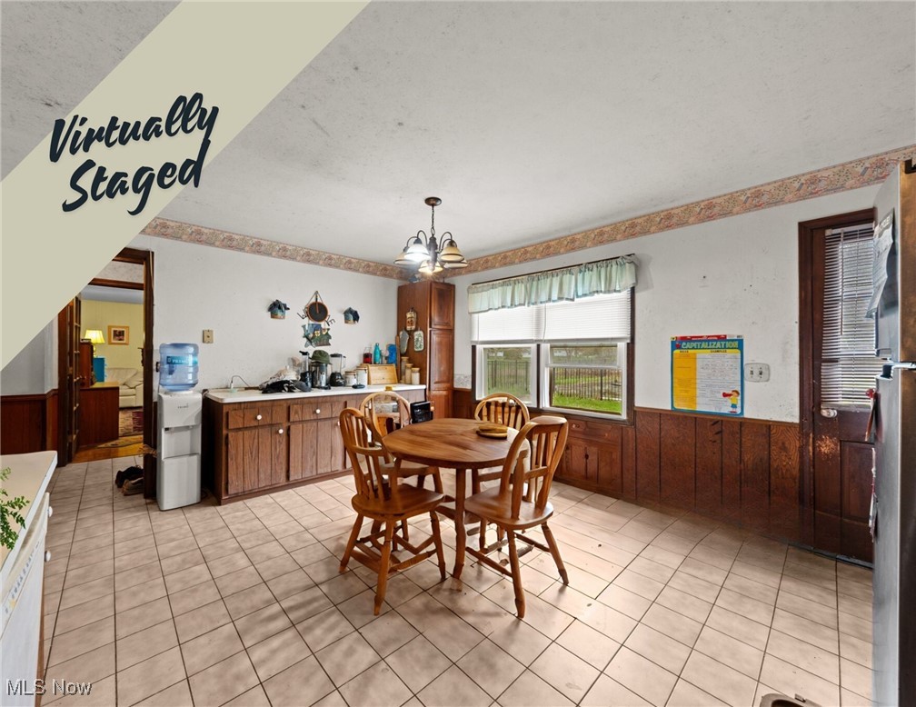 Dining area with a round wooden table, tiled floor, and adjoining kitchen; virtually staged.