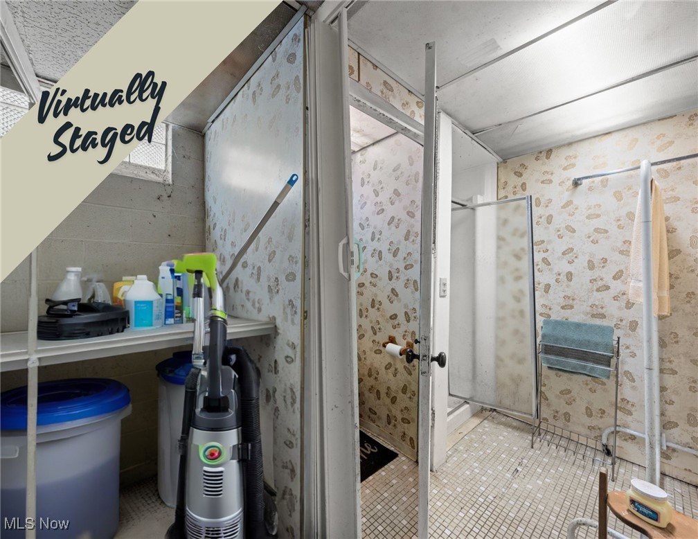 Bathroom with a tiled shower, patterned wallpaper, towel rack, and cleaning supplies.