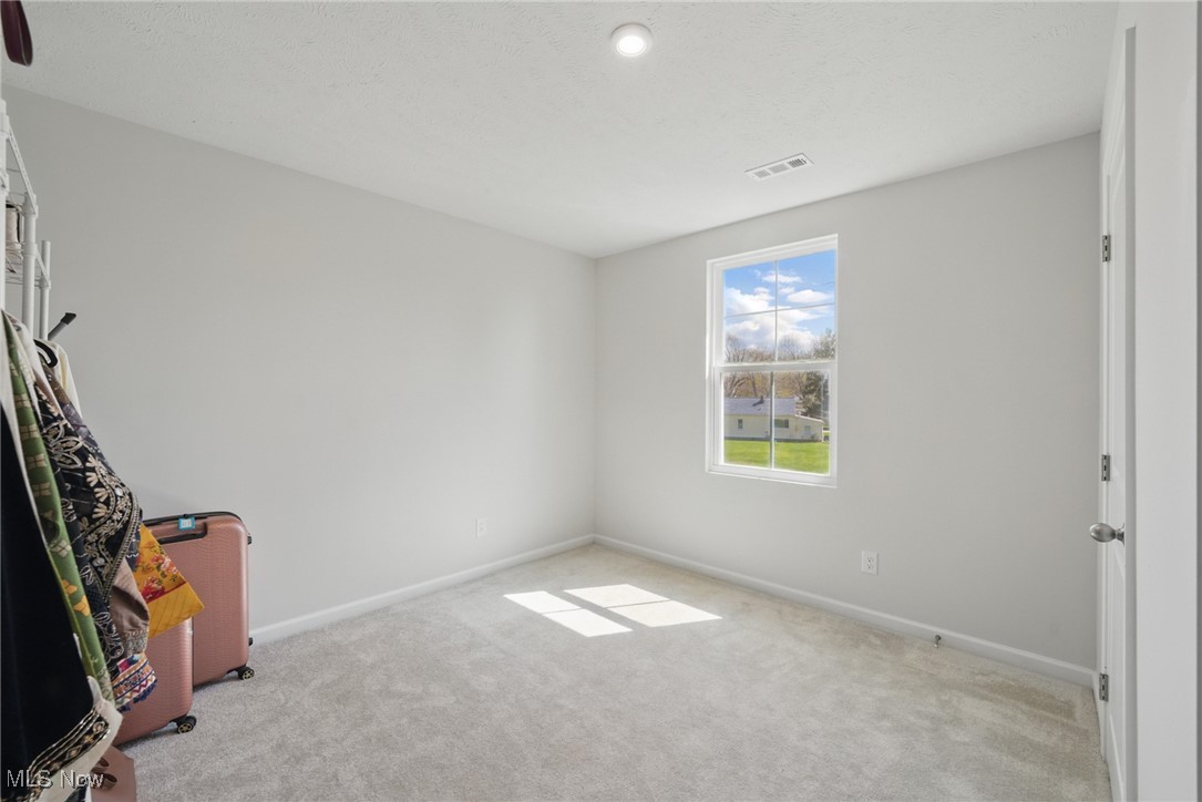 Mostly empty bedroom with beige carpet, a sunlit window, and clothing and luggage at the left edge.