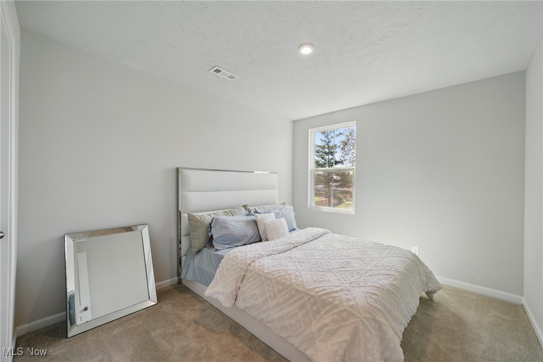 Staged bedroom with pale patterned bedding, a tall upholstered headboard, and a large leaning floor mirror.