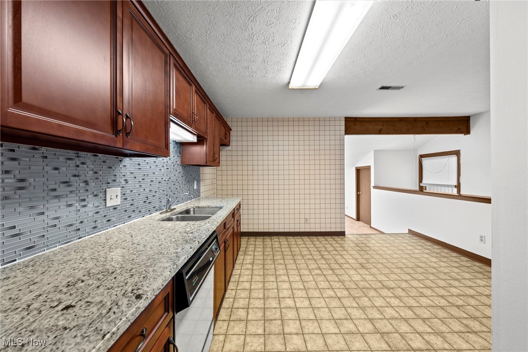 Empty kitchen with dark wood cabinets, granite counters, gray mosaic backsplash, and tan tile flooring.
