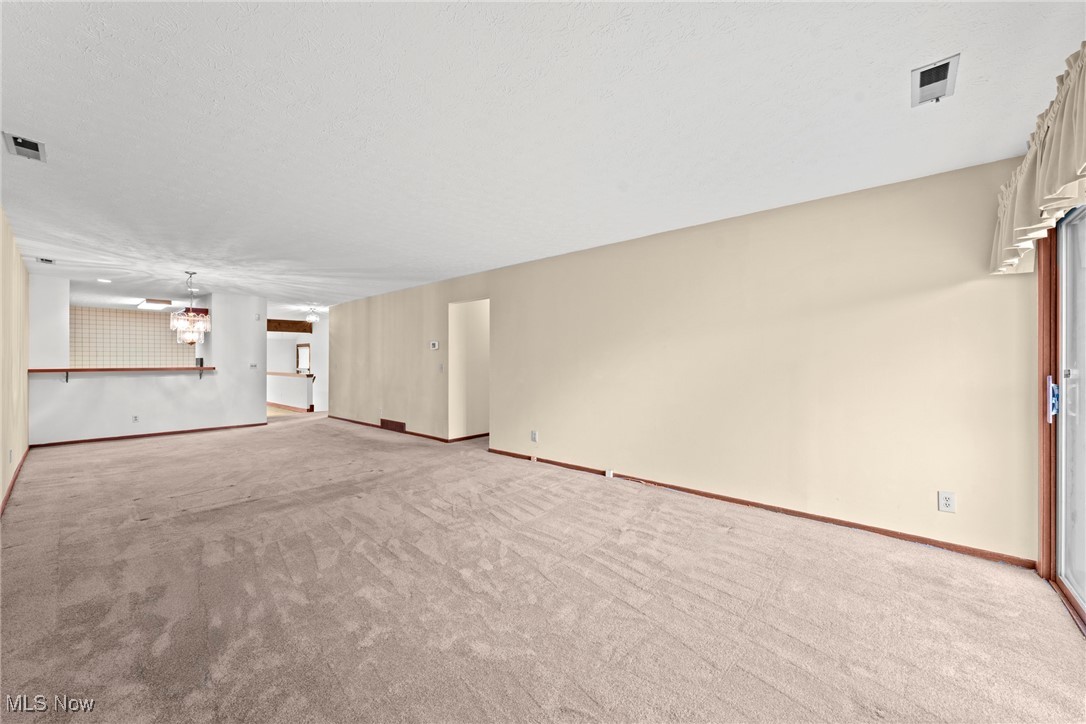 Empty carpeted living room opening to a tiled kitchen, with a sliding glass door and cream curtains.