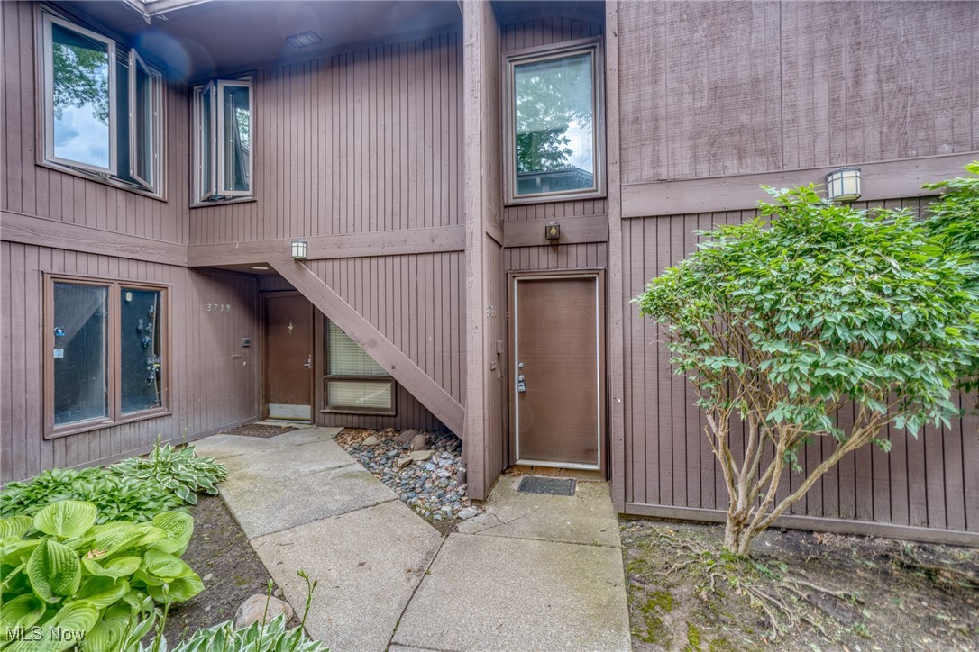 Brown-sided residential building entrance with two doors, upper windows, and landscaped shrubs.