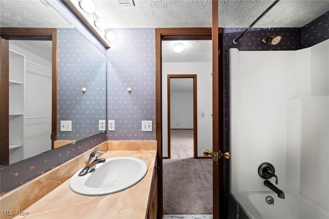 Bathroom with a marble-look vanity, oval sink, and tub-shower beside patterned wallpaper.