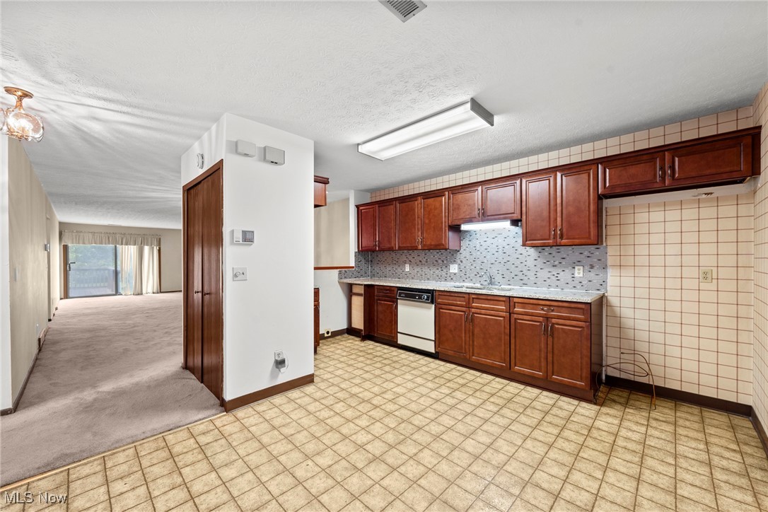 Kitchen with dark wood cabinets, tiled floor and backsplash, opening to empty carpeted living area.