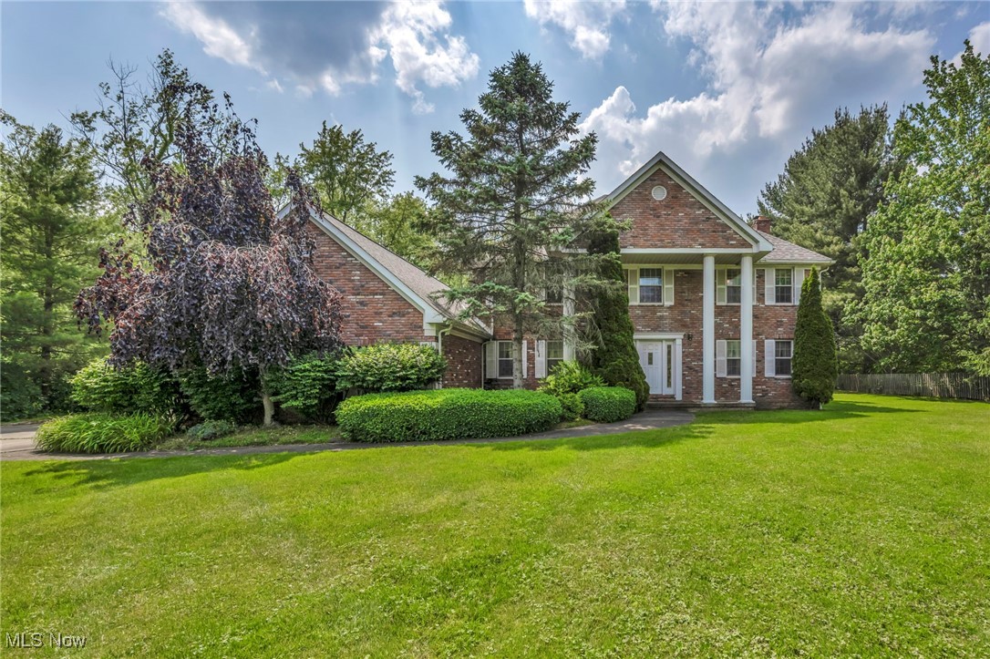 Two-story red-brick home with a columned entry, mature trees, clipped shrubs, and a broad green lawn.