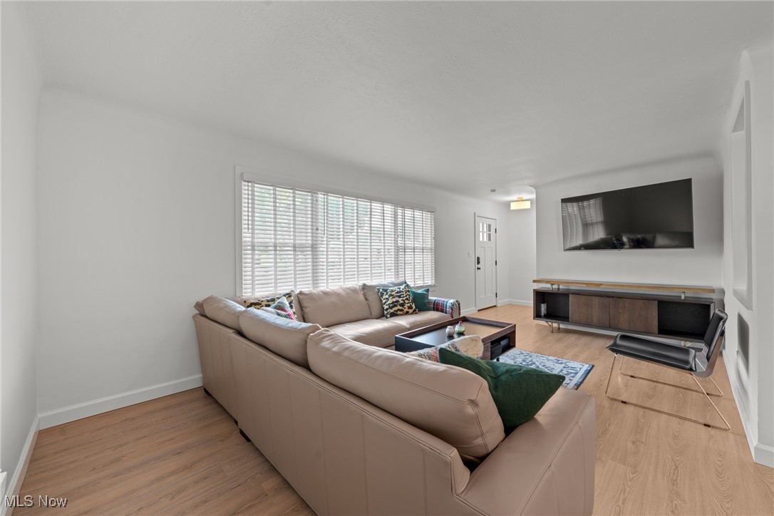 Living room with a beige sectional, wide window blinds, wood floors, and wall-mounted television.