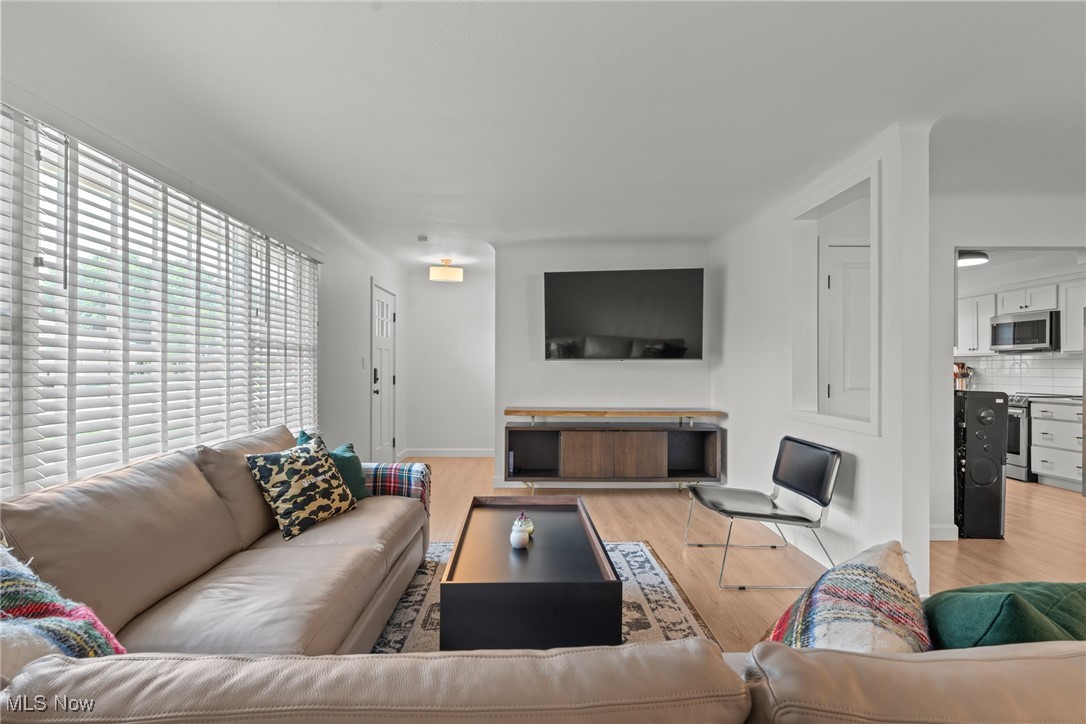 Living room with beige leather sofas, wide windows with blinds, and a wall-mounted television.