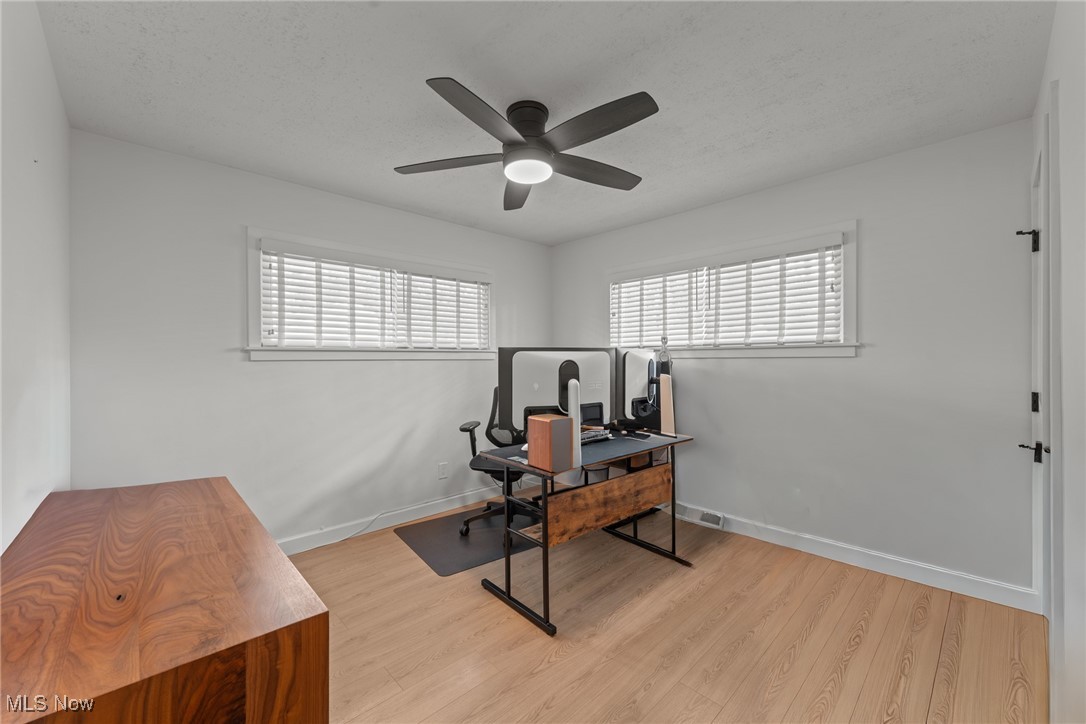 Home office with light wood floors, two high windows, a ceiling fan, and a desk with computer monitors.