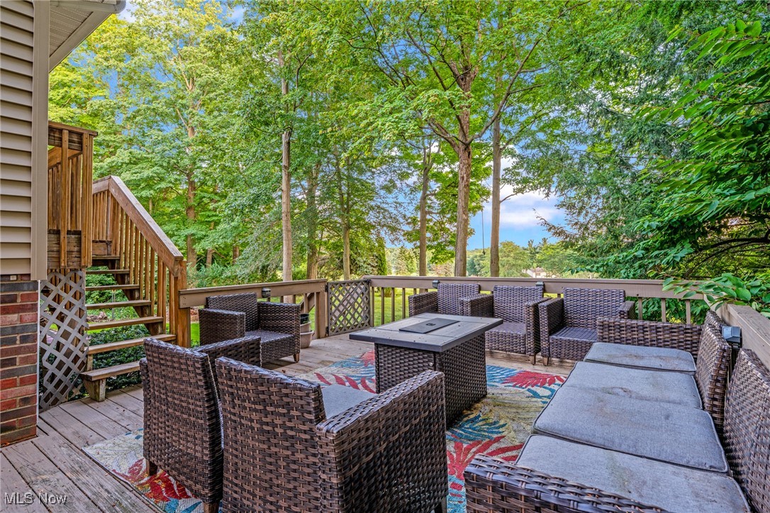 Furnished wooden deck with wicker seating and lounge chairs, surrounded by mature leafy trees.