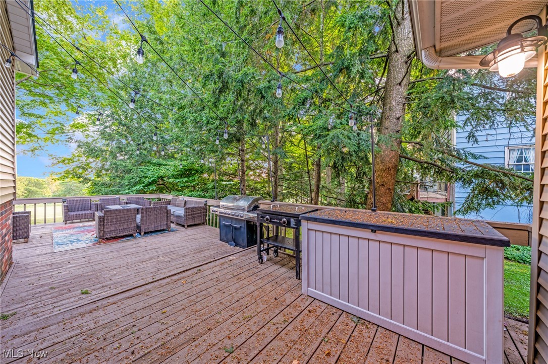 Wooden deck with wicker seating, a grill, and string lights beneath leafy trees.