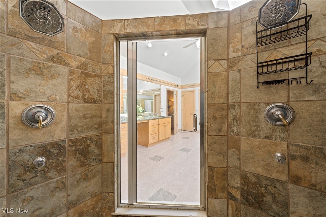 Stone-tiled shower with metal fixtures and a view through the glass door to a wood vanity.