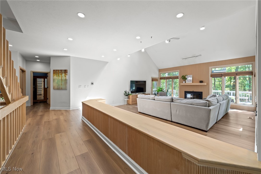 Living room with a vaulted ceiling, gray sectional, wood-paneled fireplace wall, and large windows overlooking trees.