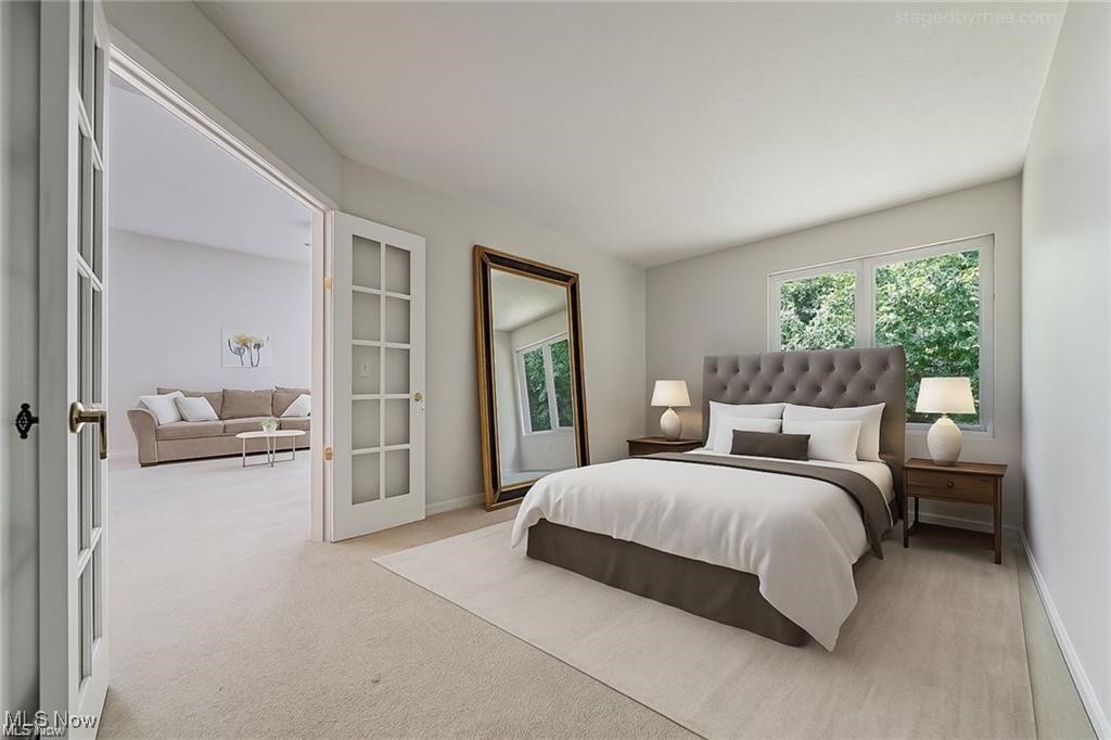 Staged bedroom with a tufted gray headboard, large floor mirror, and open doors to a sitting room.