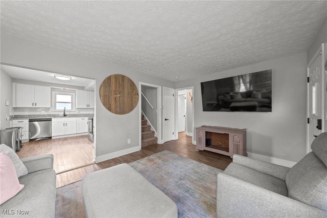 Living room opening to a white kitchen, with gray sofas, a wall-mounted TV, and round wooden wall art.