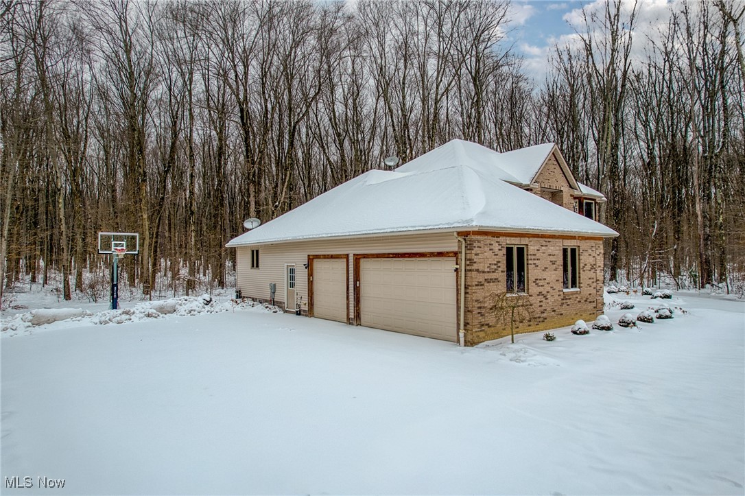 Snow-covered home with a two-door garage beside a basketball hoop and leafless woods.