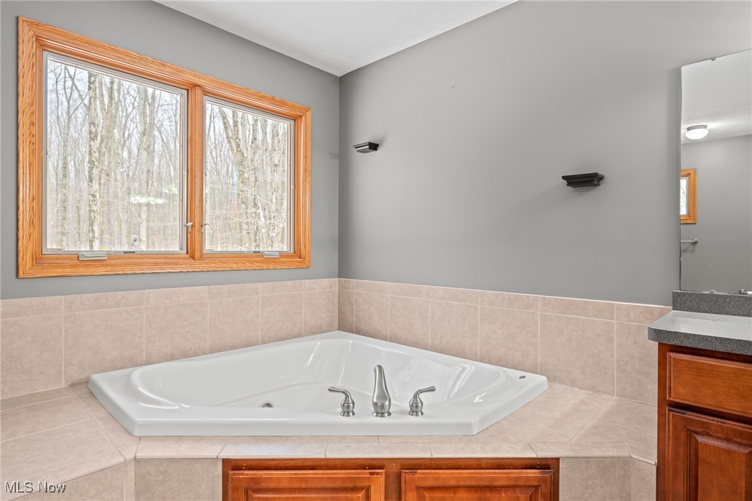 Bathroom with a corner whirlpool tub, beige tile, wood cabinetry, and windows overlooking leafless trees.