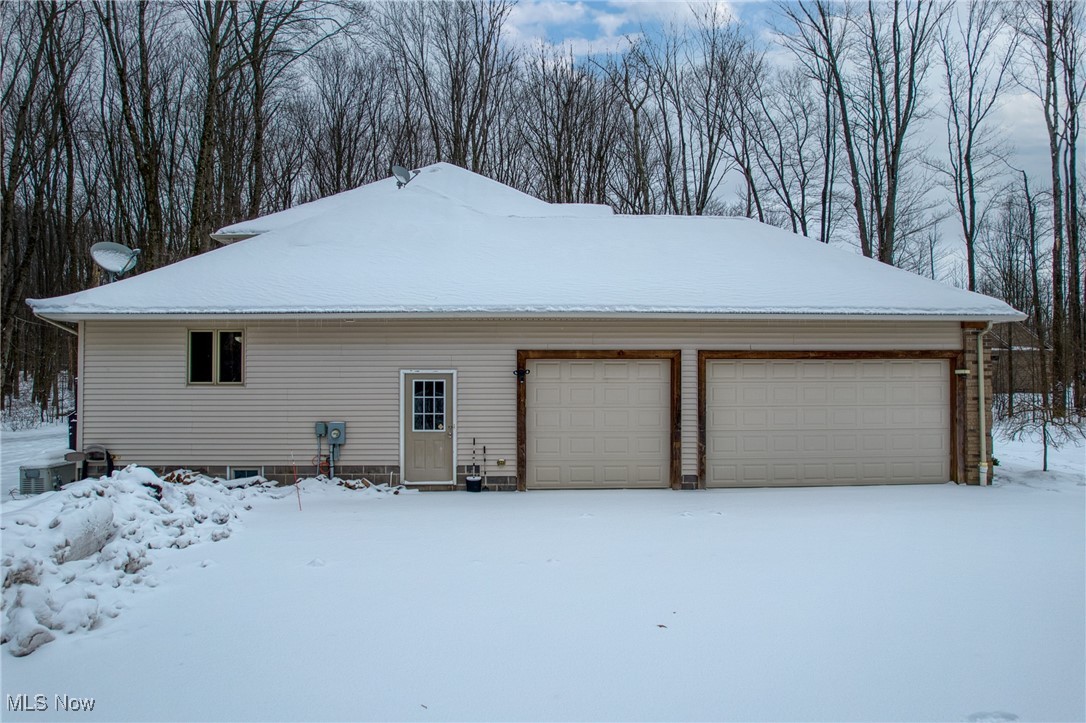 Snow-covered house exterior with two garage doors and leafless woods behind it.