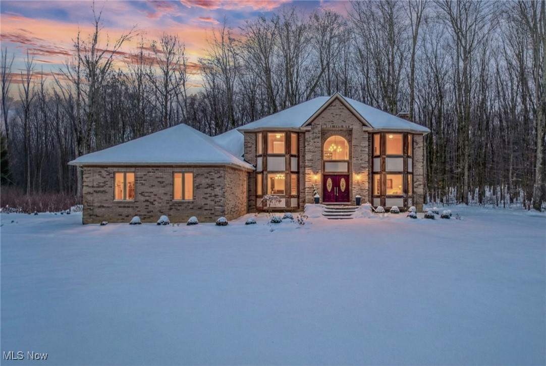 Brick two-story home with red double doors, glowing windows, and snow-covered grounds at dusk.