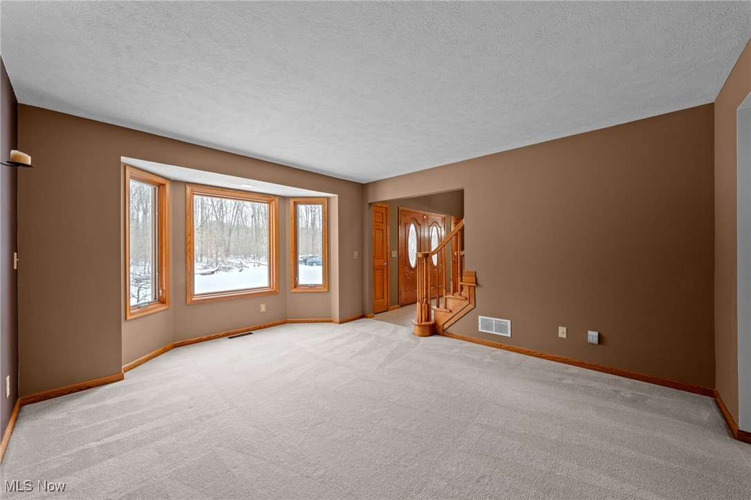 Empty living room with bay windows overlooking snowy woods and a wood staircase by the entry.