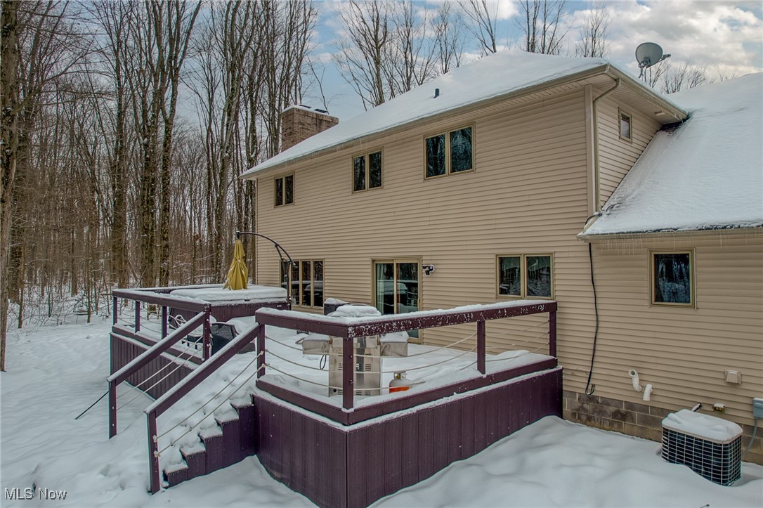 Rear of a beige two-story home with a snow-covered deck and hot tub amid winter woods.