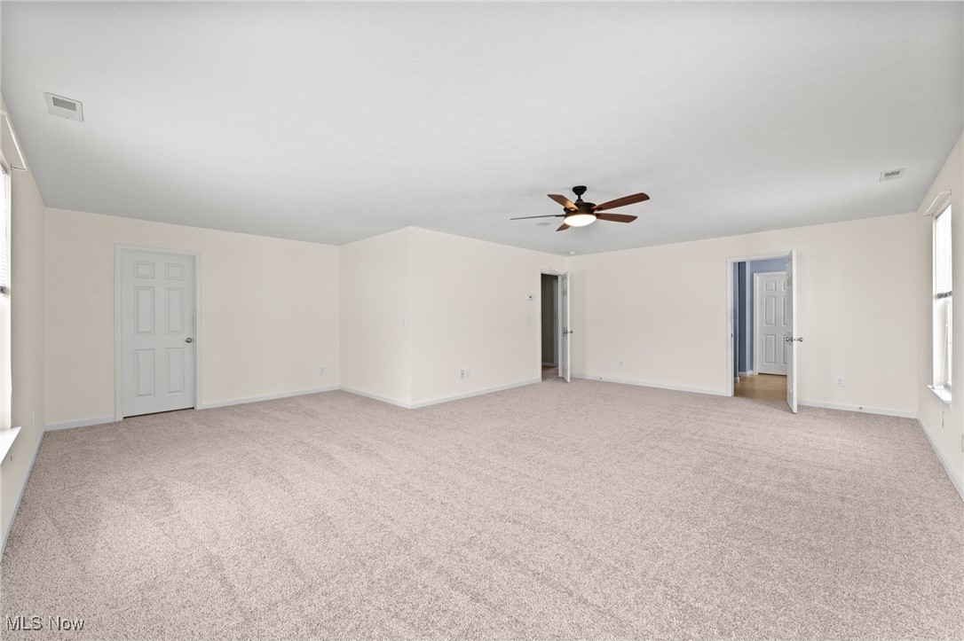 Empty carpeted room with a ceiling fan, two open doorways, a closed door, and windows.