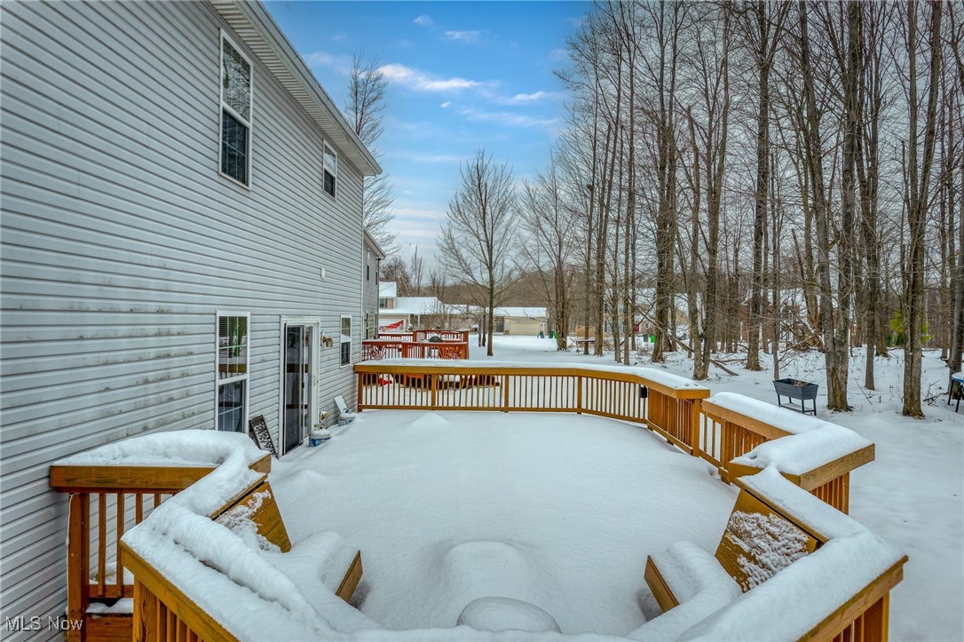 Snow-covered wooden deck beside a gray-sided house, overlooking leafless woods.